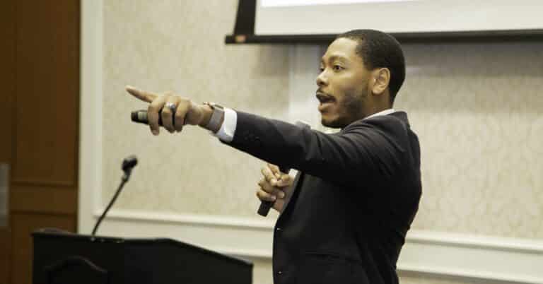 photo of Deion Jordan, a young Black man with short cropped dark hair in a black suit and white shirt, pointing out at the audience during a convening speech