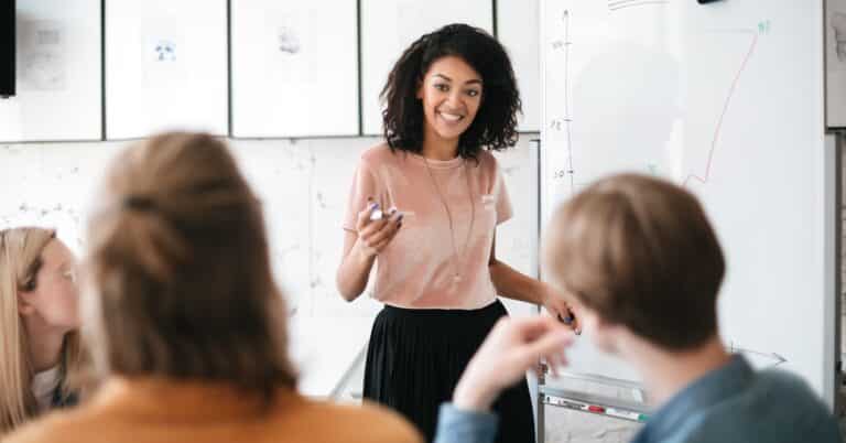 Black woman in pink top and black skirt interacting with colleagues in front of a white board